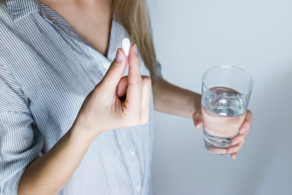 woman in blue shirt holds white pill in right hand and glass of water in left hand
