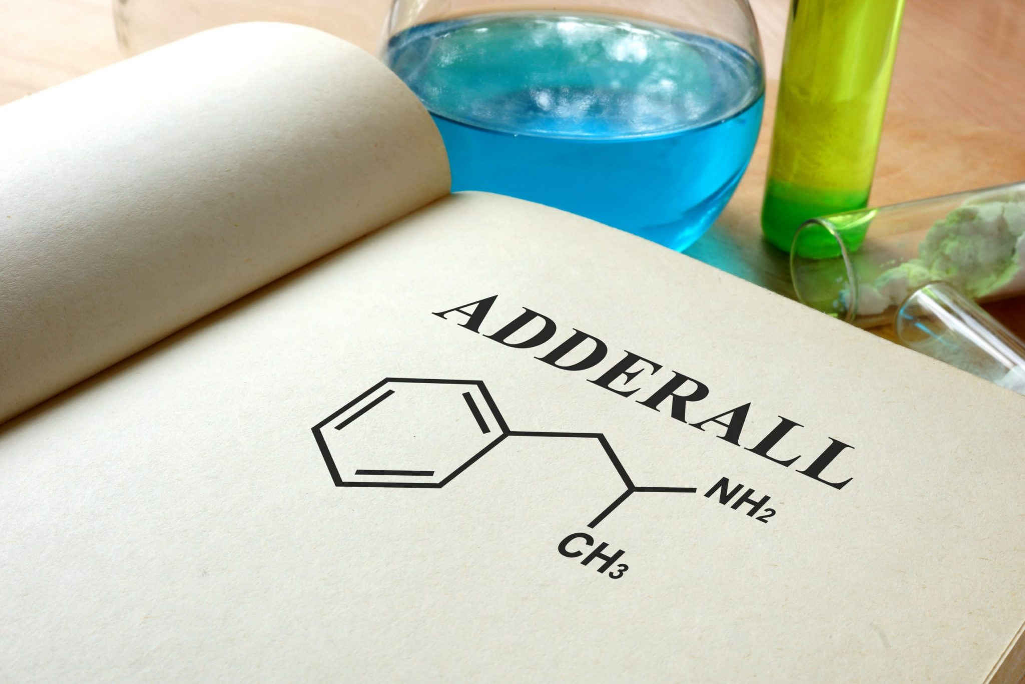 Book open to the word "ADDERALL" written over an illustration of the chemical makeup of the drug. Test tubes and a flask are partially visible in the background.