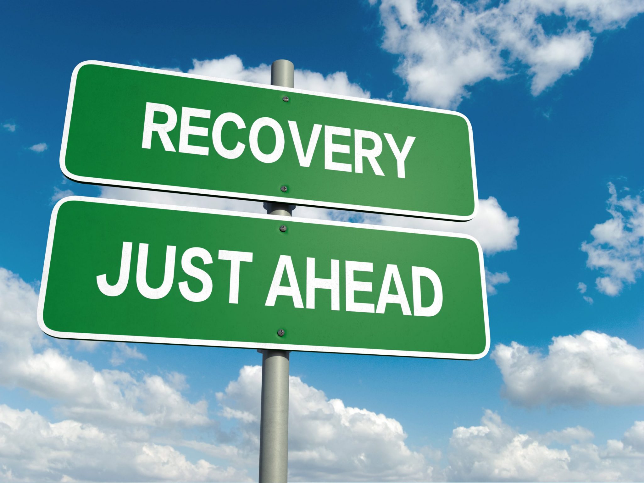 A green road sign with white lettering that says "Recovery Just Ahead". The sign is on a pole and set against a cheery blue sky dotted with puffy white clouds.