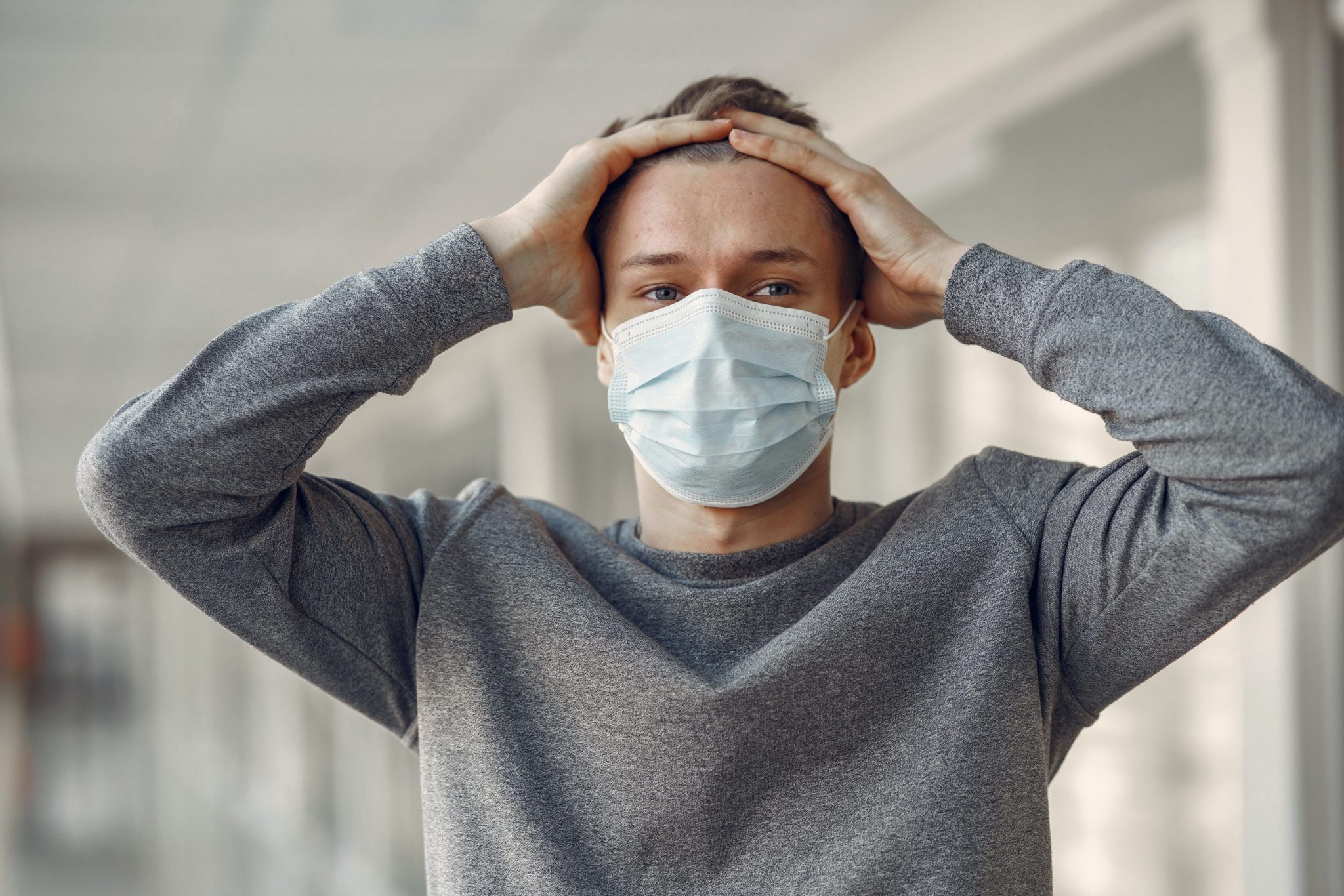 Young man wearing surgical mask, hands on head in a frustrated manner because he is coping with coronavirus stress.